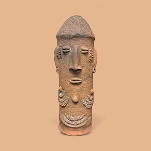 May include: A brown, cylindrical ceramic sculpture of a stylized human figure. The sculpture features a detailed face with closed eyes, a small mouth, and decorative ear and chest elements. The head is topped with a textured, conical shape.