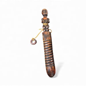 Puede incluir: Un artefacto de madera tallada con una figura humana estilizada en la parte superior de un cuerpo cilíndrico. La pieza marrón oscuro tiene un diseño acanalado y un pequeño anillo de madera sujeto con un cordón. La figura tiene los brazos cruzados.