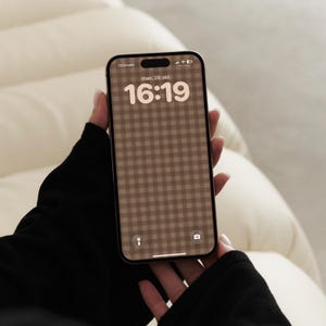 May include: A smartphone with a brown and beige gingham patterned wallpaper on the screen. The display shows the time 16:19, the date man. 20. okt., a flashlight icon, and a camera icon. The device has a black frame and a dynamic island cutout.
