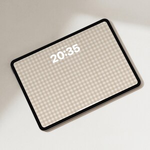Pode incluir: Um tablet com moldura preta exibindo um padr&atilde;o xadrez vichy bege e branco na tela. O rel&oacute;gio digital no visor mostra a hora 20:35 e a data sen. 30. nov. O dispositivo est&aacute; posicionado em &acirc;ngulo sobre um fundo liso e claro.