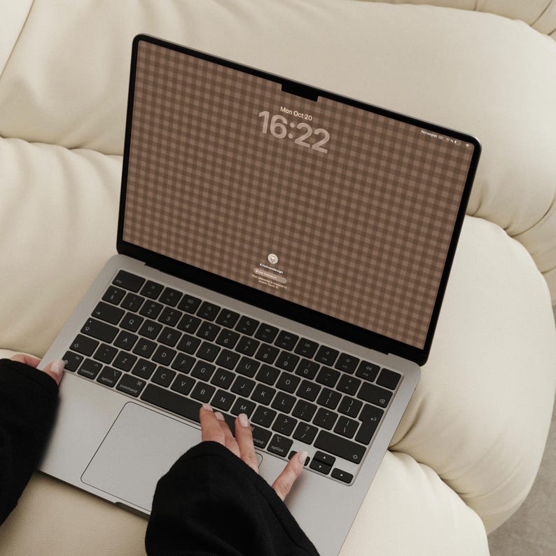 May include: A silver laptop with a black keyboard and a screen displaying a brown and white gingham pattern wallpaper. The screen shows the date Mon Oct 20 and the time 16:22 in a bold, light-coloured font centred at the top of the display.