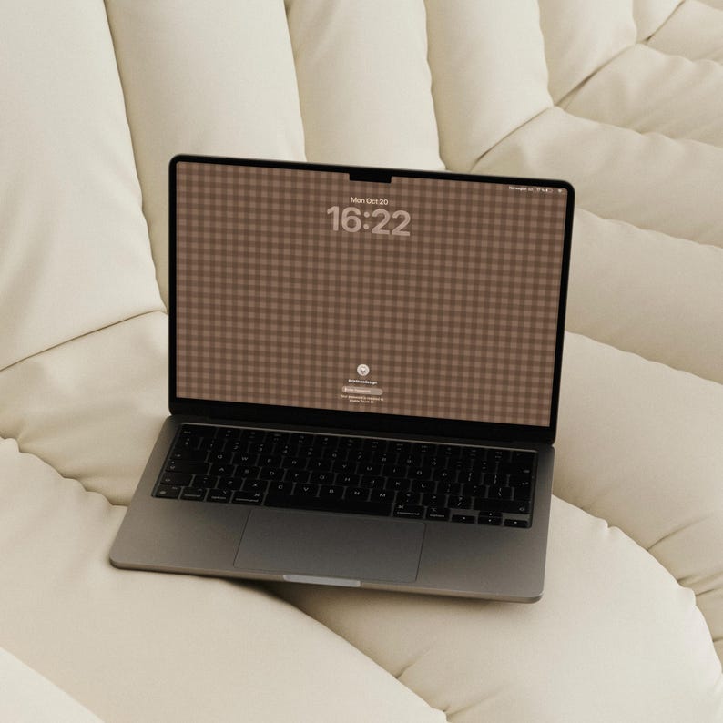 May include: A dark grey laptop with a black keyboard and a screen displaying a brown gingham pattern, the time 16:22, and the date Mon Oct 20. The laptop features a notch at the top centre of the display and a large trackpad below the keys.