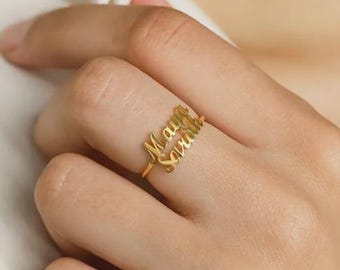 May include: A gold-colored ring with the names "Maya" and "Sarah" in a cursive font. The ring is worn on a finger, showcasing the delicate design and personalized inscription. The ring is a piece of jewelry.