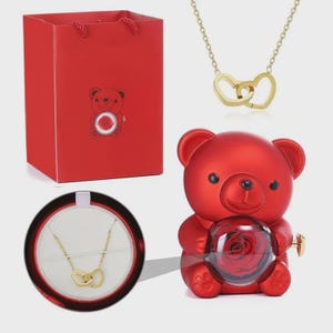 May include: A red gift bag and a red teddy bear-shaped container holding a preserved rose. A gold necklace with two interlocking heart pendants is also shown. The necklace is displayed in a round black box with a white interior.