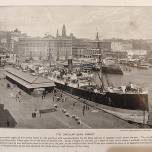 Op de afbeelding: Een vintage zwart-witfoto van Circular Quay in Sydney, Australië. Een groot schip ligt aangemeerd aan de pier, met gebouwen en andere schepen op de achtergrond. Mensen lopen op de kade.