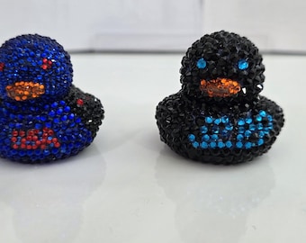 May include: Two bejeweled duck figurines. One is blue with red and orange accents, the other is black with blue and orange accents. The ducks are covered in small, sparkling stones, giving them a luxurious appearance. They are sitting on a white surface.