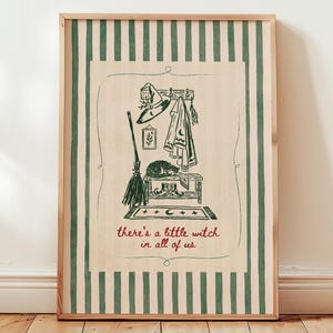 May include: A framed wall art print featuring dark green vertical stripes on a cream background. The central illustration depicts a broom, a hat, a cloak, and a cat on a bench, with the red text "there's a little witch in all of us" below.