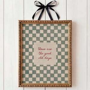 May include: Framed artwork with a gold beaded frame and a black ribbon. The artwork features a green and white checkered pattern with the text "these are the good old days" in red. The frame is hanging on a white wooden wall.