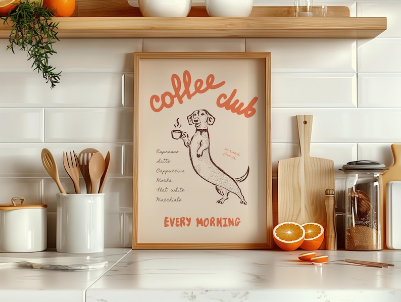 Op de afbeelding: Een ingelijste print met een teckel die een koffiekopje vasthoudt, met de woorden "Coffee Club" in oranje script. De print geeft koffiedranken weer en de woorden "Every Morning" in het oranje. De print staat in een keuken.