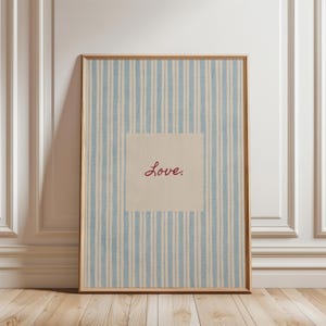 May include: Framed artwork with a light blue and white striped background. A square in the center features the word "Love" in a cursive, reddish-brown font. The frame is a light brown color.