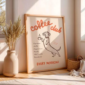 Op de afbeelding: Een ingelijste print met de woorden "coffee club" in rode letters boven een cartoon teckel die een kopje koffie vasthoudt. De print bevat een lijst met koffiedranken en de woorden "every morning". De lijst is van licht hout.