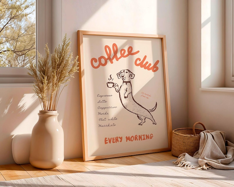 Op de afbeelding: Een ingelijste print met de woorden "coffee club" in rode letters boven een cartoon teckel die een kopje koffie vasthoudt. De print bevat een lijst met koffiedranken en de woorden "every morning". De lijst is van licht hout.