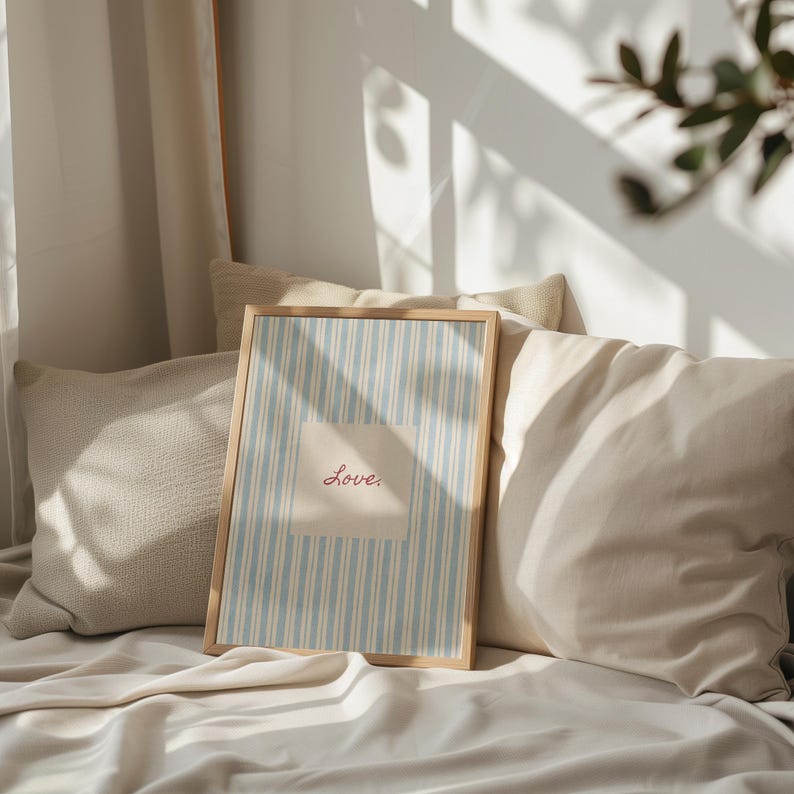 May include: A framed print with a light blue and white striped background and the word "Love" in red script. The print is in a light wood frame and rests on a bed with beige pillows and a white sheet.