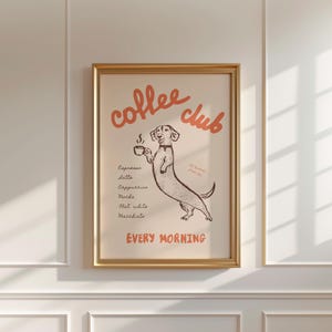 Op de afbeelding: Een ingelijste print met een beige achtergrond en de woorden "coffee club" in oranje script. Een cartoon teckel hond houdt een kopje koffie vast. Onder de hond staan namen van koffiedranken. De woorden "every morning" staan onderaan.