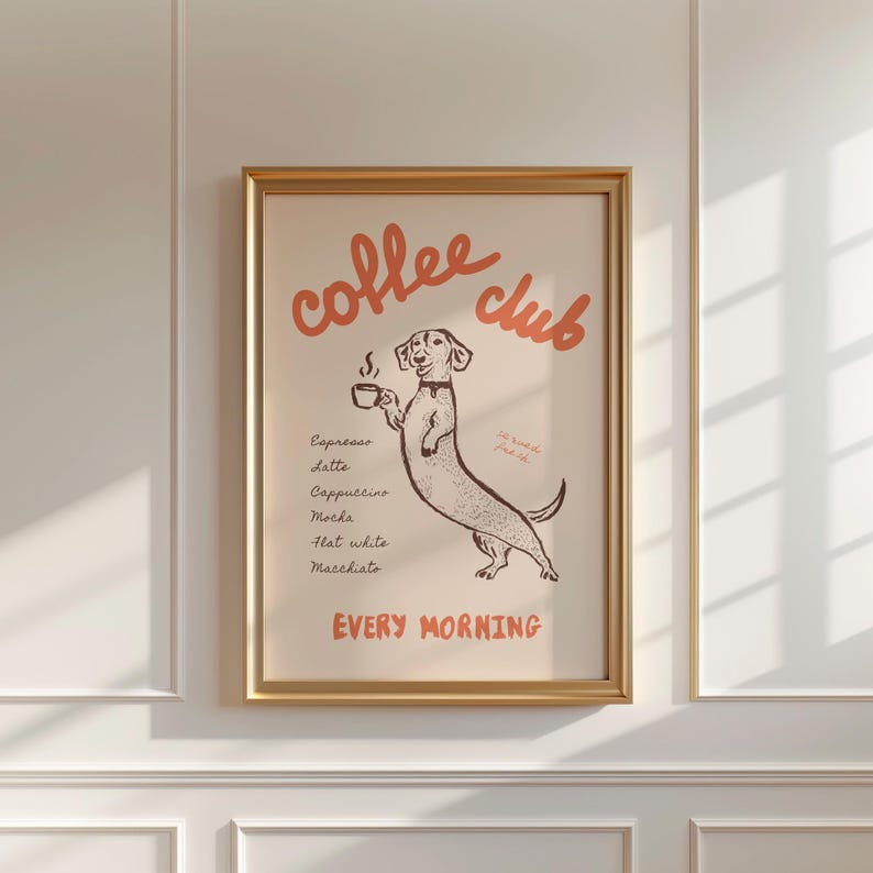 Op de afbeelding: Een ingelijste print met een beige achtergrond en de woorden "coffee club" in oranje script. Een cartoon teckel hond houdt een kopje koffie vast. Onder de hond staan namen van koffiedranken. De woorden "every morning" staan onderaan.