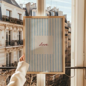 May include: A framed print with a light wood frame. The print features a blue and white striped background with a square off-white center. The word "Love" is written in red script within the square.