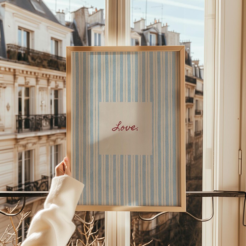 May include: A framed print with a light wood frame. The print features a blue and white striped background with a square off-white center. The word "Love" is written in red script within the square.