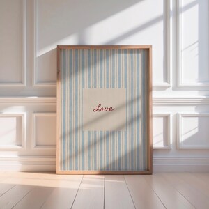 May include: A framed print featuring a light blue and white striped background with a beige square in the center. The word "Love" is written in red cursive script within the square. The frame is made of light brown wood.