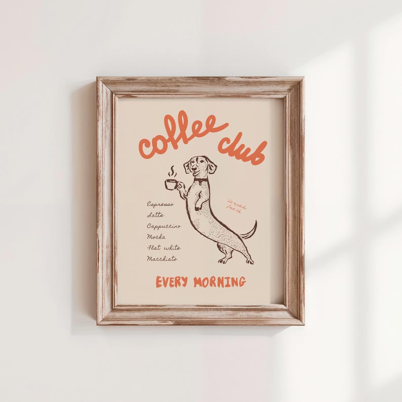 Op de afbeelding: Ingelijste print met een illustratie van een teckel die een kopje koffie vasthoudt. De woorden "coffee club" staan in oranje letters boven de hond, met een lijst met koffiedranken eronder. De zin "every morning" staat onderaan. De lijst is lichtbruin.