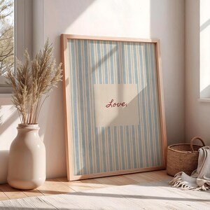 May include: A framed artwork with a light blue and white striped background and the word "Love" in red script. A beige vase with dried plants sits next to the artwork. A woven basket and a folded blanket are also in the image.