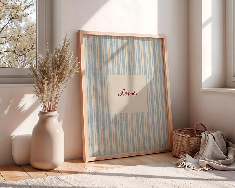 May include: A framed artwork with a light blue and white striped background and the word "Love" in red script. A beige vase with dried plants sits next to the artwork. A woven basket and a folded blanket are also in the image.