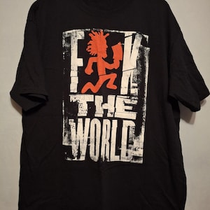 Może przedstawiać: Czarny t-shirt z postarzaną biało-pomarańczową grafiką. Grafika przedstawia biegnącą postać trzymającą siekierę, z napisem "F**K THE WORLD" pogrubioną czcionką. Koszulka wisi na czarnym wieszaku.