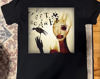 Pode incluir: T-shirt preto com um design gráfico. O design inclui as palavras "GET SCARED", um corvo em uma mão e um retrato surrealista de uma mulher com pele rachada e detalhes em vermelho.