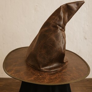 Peut inclure: Un chapeau de sorcière marron avec un large bord et une couronne pointue. Le chapeau a un aspect texturé semblable à du cuir et une couleur marron foncé. Le bord a une bordure plus foncée. Le chapeau est présenté sur une surface noire.