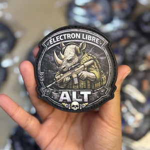 Op de afbeelding: Een zwart en grijs embleem met een neushoorn in een gele vest die een geweer vasthoudt. Het embleem heeft de tekst "ÉLECTRON LIBRE" bovenaan en "ALT" onderaan, met een schedelafbeelding.
