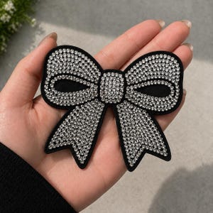 May include: A black bow-shaped accessory, embellished with numerous sparkling rhinestones. The bow has a solid black center and edges, with the rhinestones covering the bow's loops and tails. This decorative item could be used for clothing or accessories.