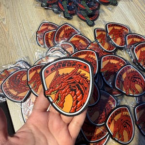 Könnte beinhalten: Eine Sammlung von Aufnähern mit einem roten Drachendesign und dem Text "Family Airsoft" auf einem schildförmigen Aufnäher. Andere Aufnäher zeigen ein schwarz-rotes Design mit dem Text "WACAT GHOSTS".