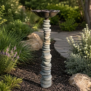 May include: A birdbath featuring a tall, slender pedestal constructed from a vertical stack of smooth, rounded gray river stones. At the top sits a dark, metallic, flower-shaped basin with scalloped edges designed to hold water for birds.