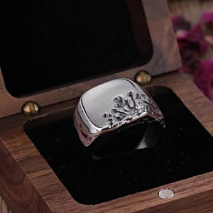 May include: A silver ring with a square face and a textured design on one side, presented in an open wooden jewellery box with a black velvet interior. The ring is the focal point, with rose petals scattered around the box.