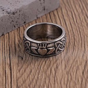 Puede incluir: Un anillo plateado con un diseño de Claddagh. El anillo presenta un motivo de corazón, corona y manos, rodeado de patrones de nudos celtas. El anillo está sobre una superficie de madera y un bloque gris.