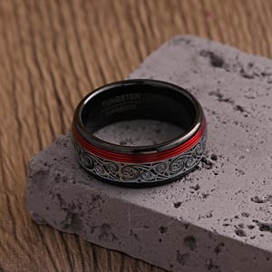 May include: A black tungsten carbide ring with a red stripe and a silver Celtic knot design. The ring is on a gray, textured surface. The ring has the words "Tungsten Carbide" engraved on the inside.
