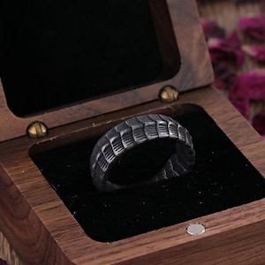 May include: A textured, dark gray ring with a geometric pattern rests inside a wooden ring box with a black velvet interior. The box is open, revealing the ring. Rose petals are scattered around the box.