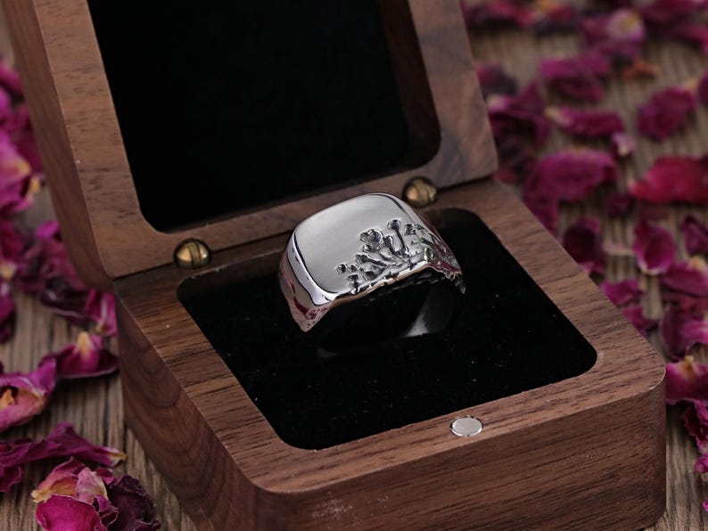 May include: A silver ring with a square face and a textured design on one side, presented in an open wooden jewellery box with a black velvet interior. The ring is the focal point, with rose petals scattered around the box.