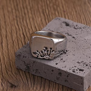 May include: A silver signet ring with a square face and a textured design. The ring sits on a grey, rectangular block, with a wooden surface in the background. The ring's design appears to be a stylized floral or organic pattern.