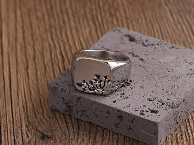 May include: A silver signet ring with a square face and a textured design. The ring sits on a grey, rectangular block, with a wooden surface in the background. The ring's design appears to be a stylized floral or organic pattern.
