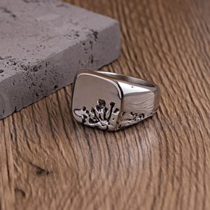 May include: A silver ring with a square face and a textured design. The ring is placed on a wooden surface beside a grey rectangular block. The ring's design resembles a stylized coral or plant-like pattern.