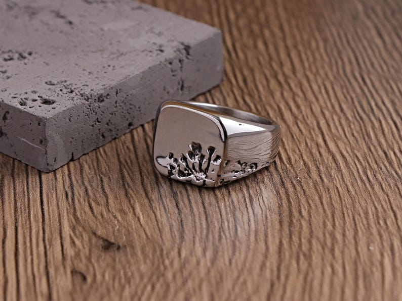 May include: A silver ring with a square face and a textured design. The ring is placed on a wooden surface beside a grey rectangular block. The ring's design resembles a stylized coral or plant-like pattern.