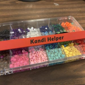 May include: A clear plastic organizer box filled with colorful beads. The beads are in various colors, including pink, blue, yellow, and orange. A red label across the top reads "Kandi Helper".