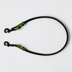Puede incluir: Una correa negra con un dise&ntilde;o de cord&oacute;n liso y redondeado. Cada extremo de la correa est&aacute; equipado con un mosquet&oacute;n giratorio de metal negro, asegurado por una pieza de conexi&oacute;n de color verde lima brillante con dos remaches negros a cada lado.