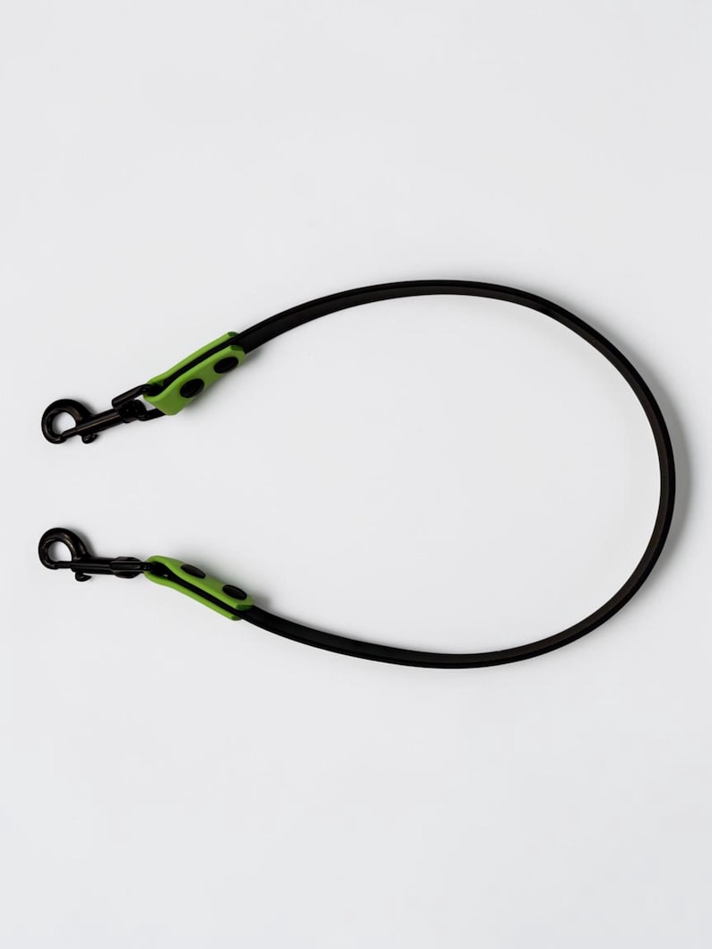 Puede incluir: Una correa negra con un dise&ntilde;o de cord&oacute;n liso y redondeado. Cada extremo de la correa est&aacute; equipado con un mosquet&oacute;n giratorio de metal negro, asegurado por una pieza de conexi&oacute;n de color verde lima brillante con dos remaches negros a cada lado.