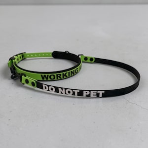 Puede incluir: Un conjunto de collar y correa en color verde lima y negro. El collar presenta el texto WORKING en negro, mientras que la correa muestra el texto DO NOT PET en blanco. Ambos art&iacute;culos est&aacute;n hechos de un material liso y flexible con hebillas negras.