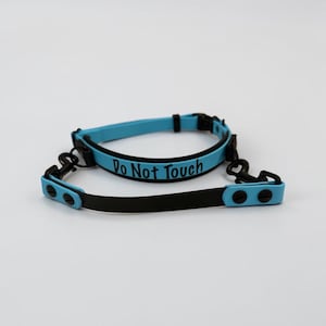 Puede incluir: Un collar azul claro con bordes y herrajes negros, que presenta el texto "Do Not Touch" en letras negras. El collar incluye una correa negra a juego con extremos azul claro, asegurada mediante remaches negros y ganchos met&aacute;licos giratorios.