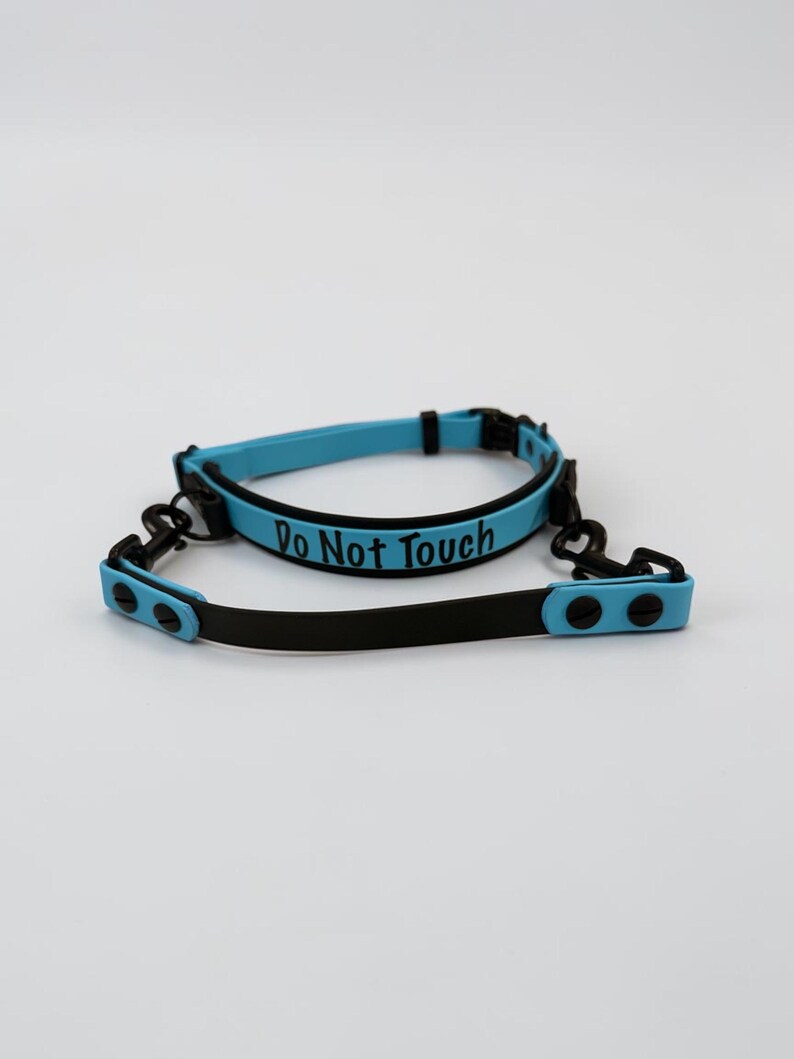 Puede incluir: Un collar azul claro con bordes y herrajes negros, que presenta el texto "Do Not Touch" en letras negras. El collar incluye una correa negra a juego con extremos azul claro, asegurada mediante remaches negros y ganchos met&aacute;licos giratorios.