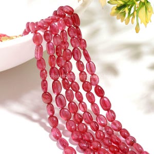 May include: A strand of smooth, oval-shaped ruby-colored gemstone beads arranged in multiple vertical rows. The beads exhibit a translucent, deep red hue with natural variations in color intensity and clarity, reflecting light across their polished surfaces.