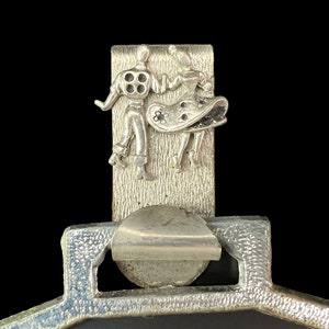 Puede incluir: Un espejo compacto de metal plateado con una tapa con bisagras. La tapa presenta un diseño en relieve de un hombre y una mujer bailando.