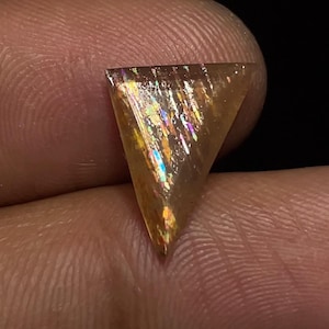 Puede incluir: Una gema triangular e iridiscente con un color base marrón dorado cálido. La gema muestra un juego de colores, con destellos de tonos arcoíris en su superficie. La piedra se sostiene entre los dedos sobre un fondo oscuro.